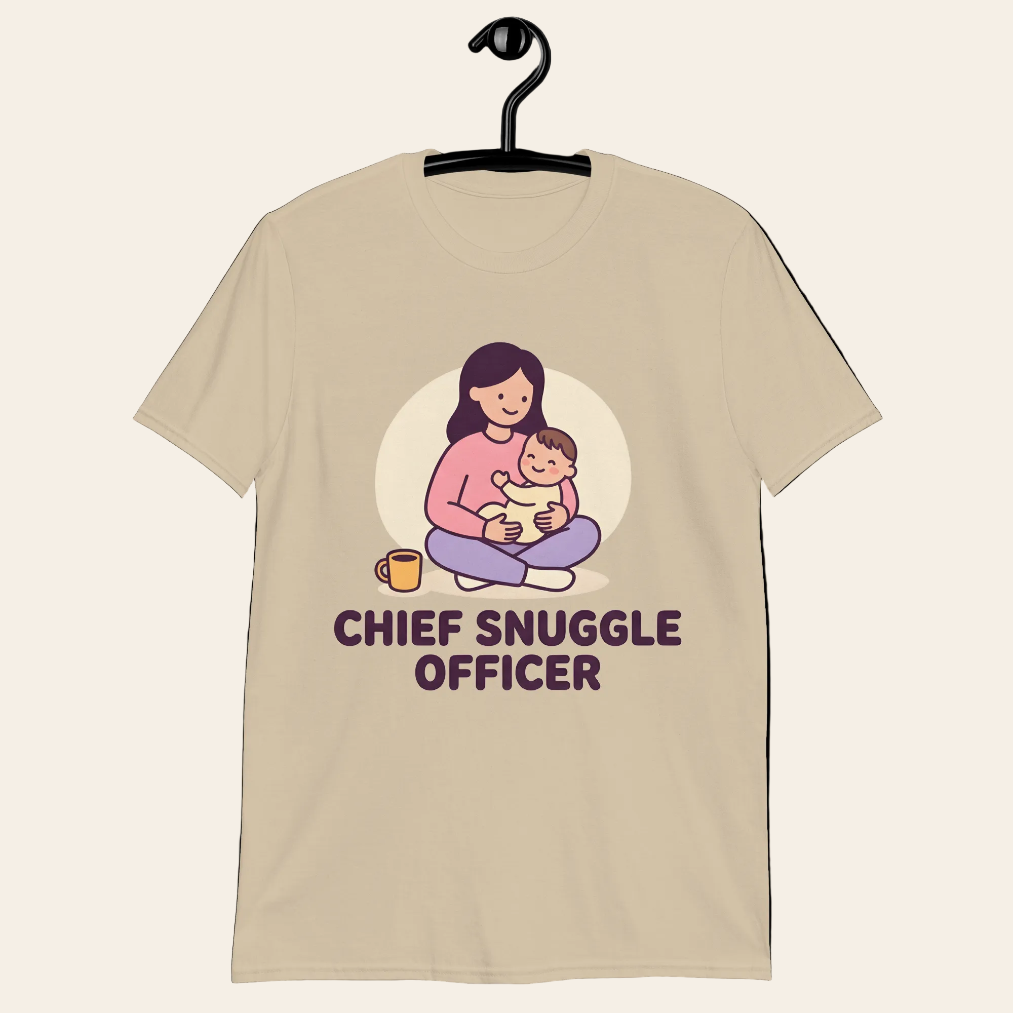 A beige short-sleeve t-shirt hanging on a black hanger, featuring a cartoon graphic of a mother holding her baby with the text 'CHIEF SNUGGLE OFFICER' underneath.